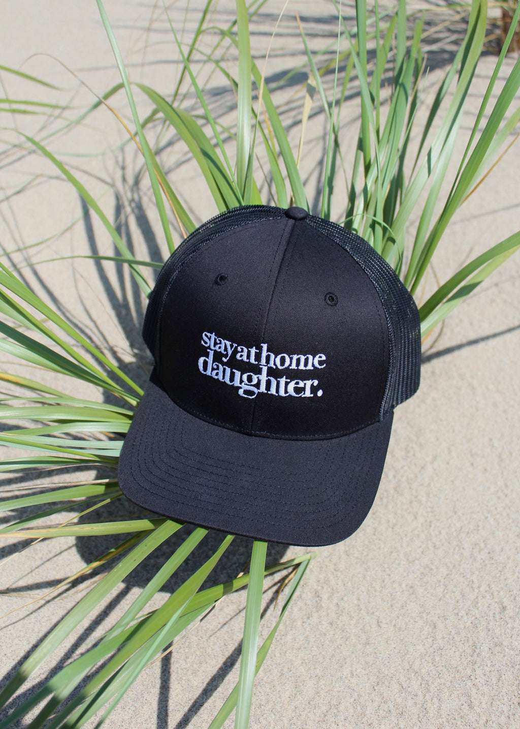 A black trucker-style cap with white embroidered text reading 'stay at home daughter.' resting on green grass over a sandy surface.