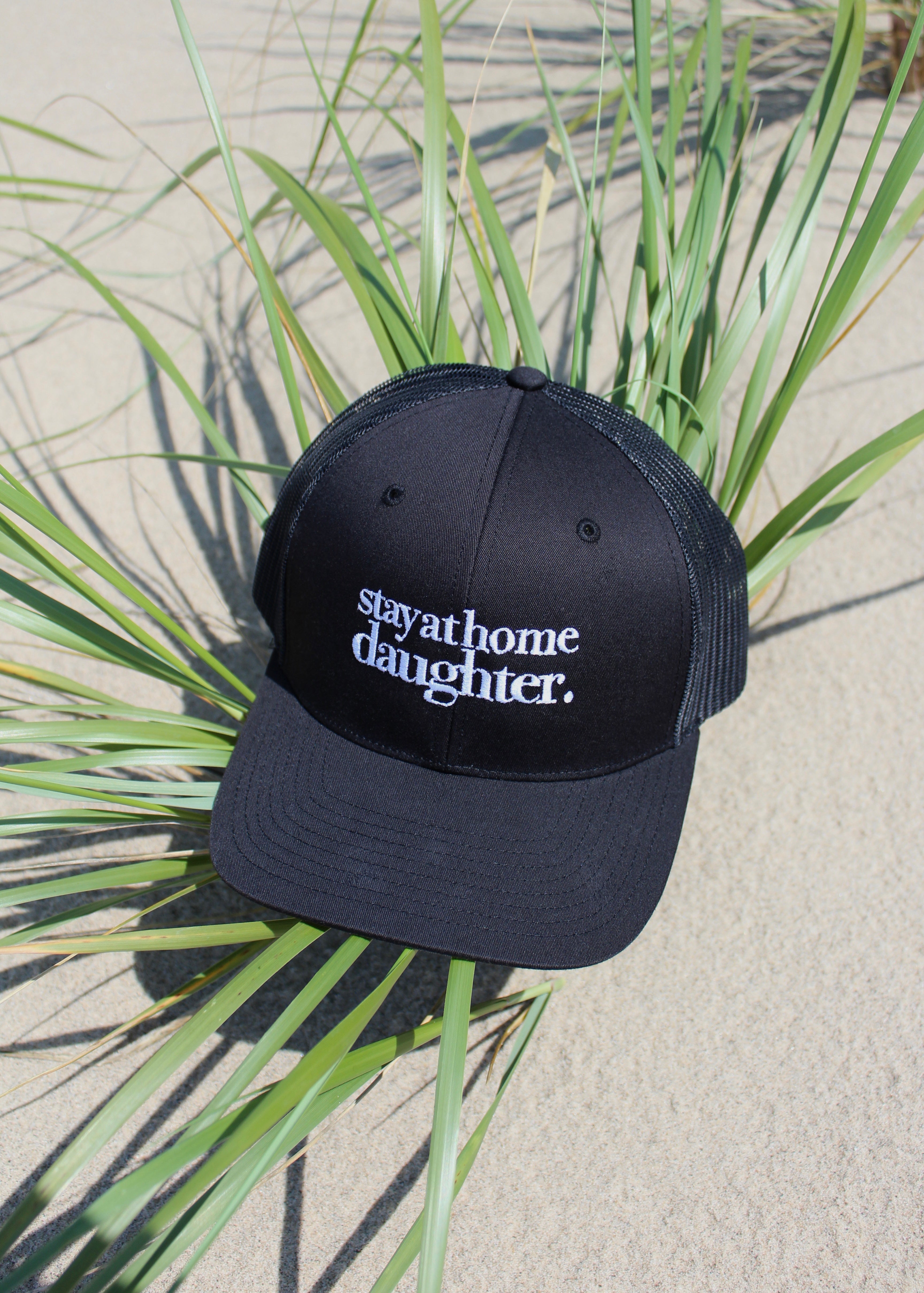 A black trucker-style cap with white embroidered text reading 'stay at home daughter.' resting on green grass over a sandy surface.