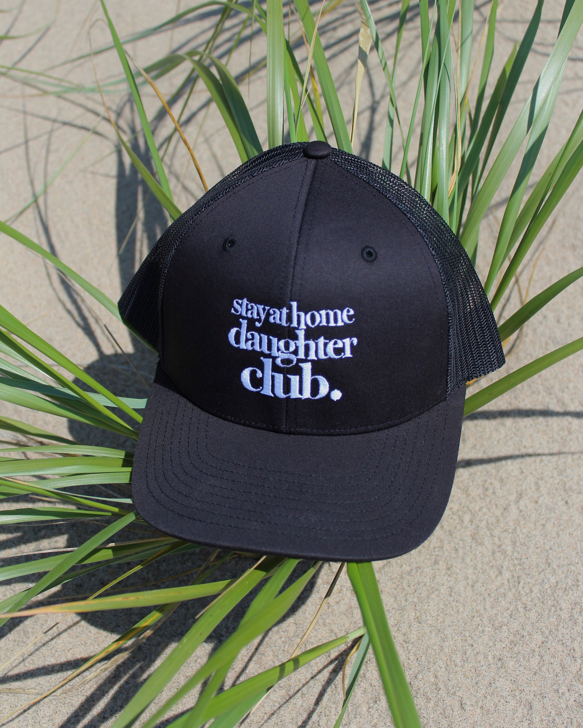 A black trucker-style baseball cap with white embroidered text reading 'stay at home daughter club.' placed on sand among green beach grass.