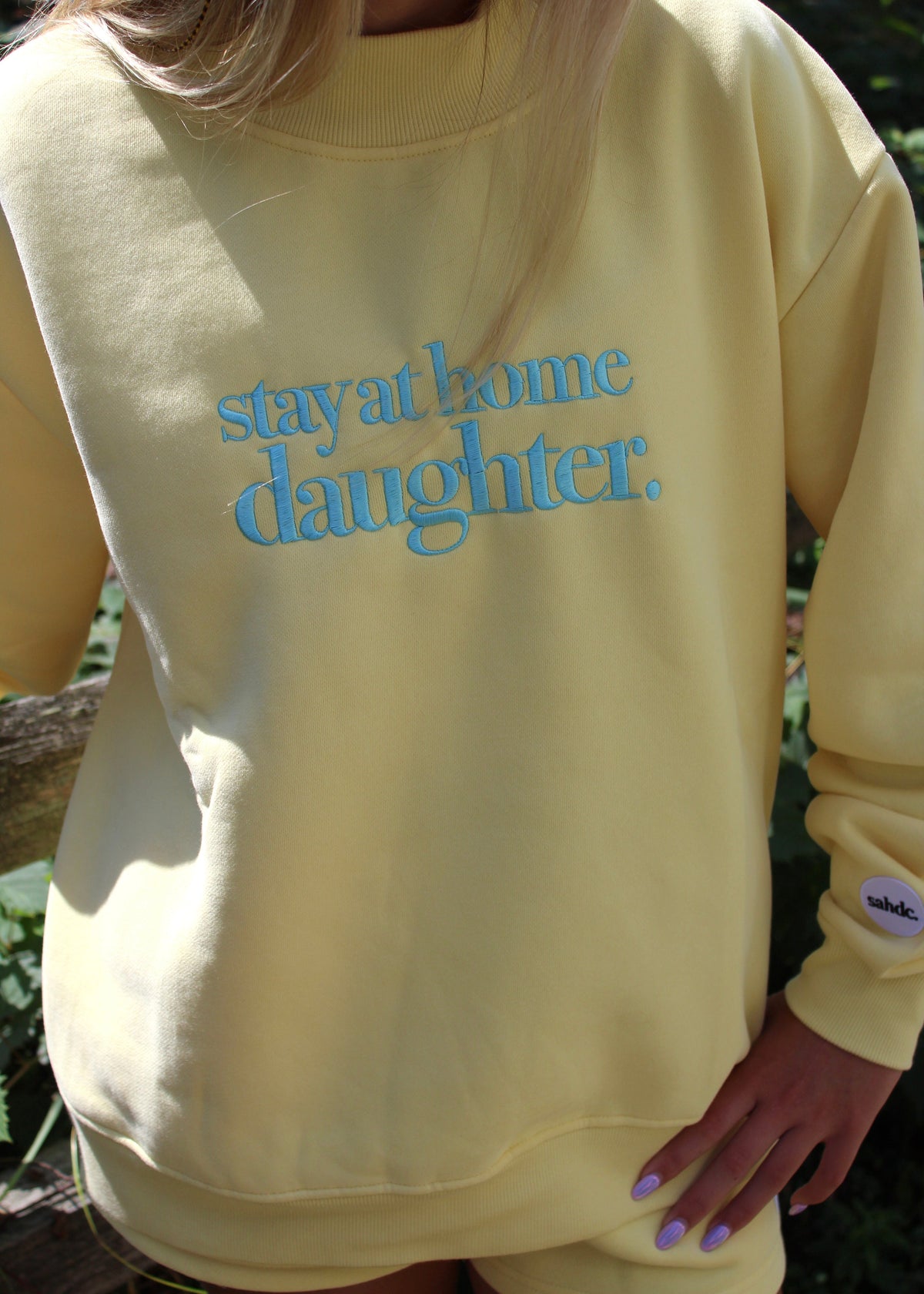A person wearing a pale yellow sweatshirt with the phrase 'stay at home daughter.' embroidered in light blue across the chest, photographed outdoors with foliage in the background.