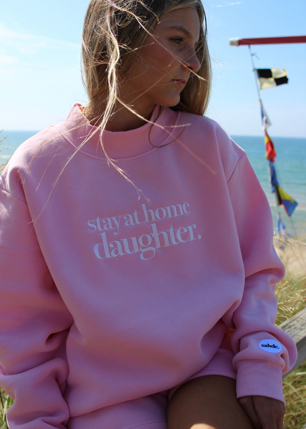 A young girl wearing a pink sweatshirt with the text 'stay at home daughter.' printed in white on the front, set against a beach background with ocean and sky visible.