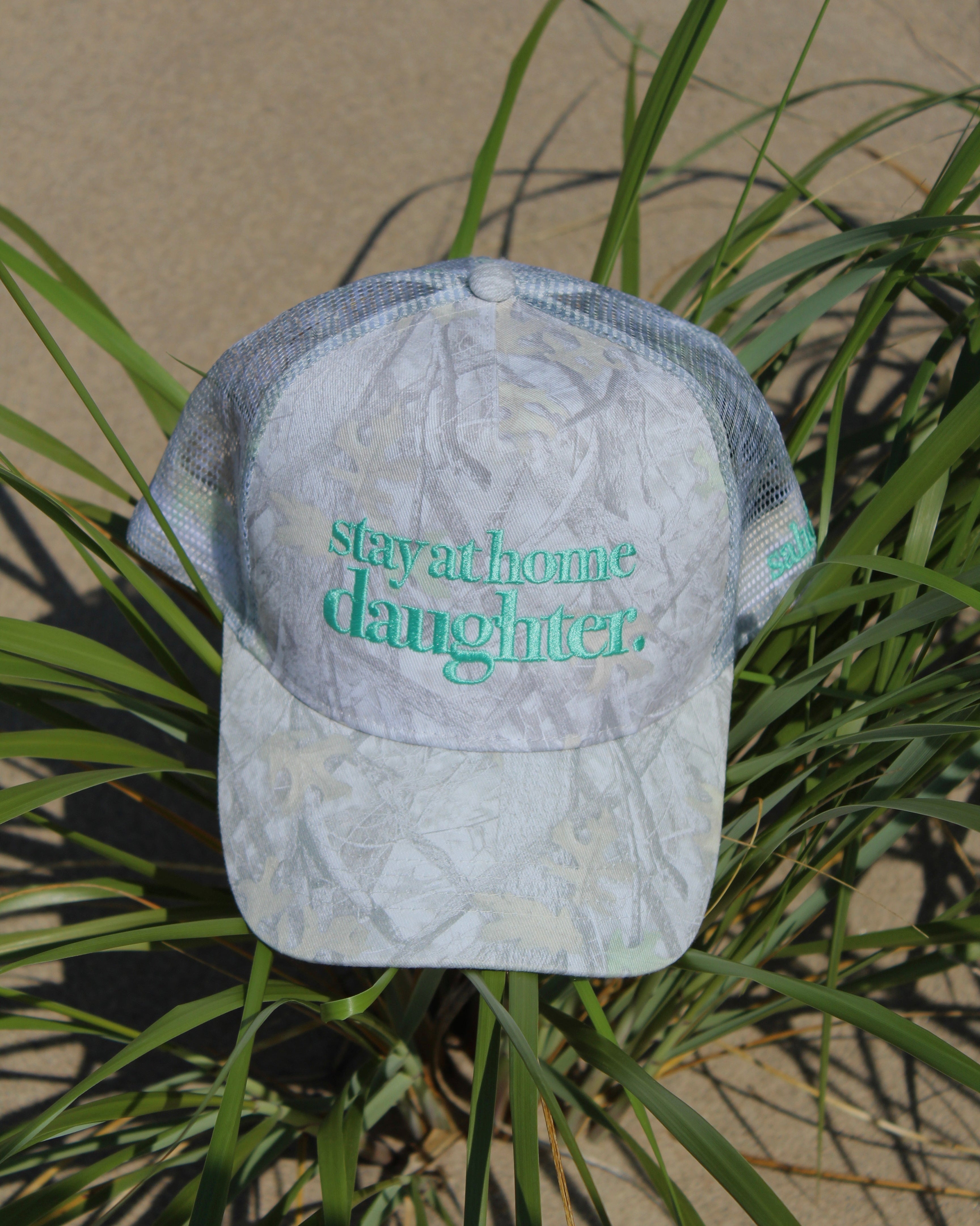 A light gray and white camouflage trucker hat with 'stay at home daughter.' embroidered in teal on the front, resting on green beach grass outdoors.