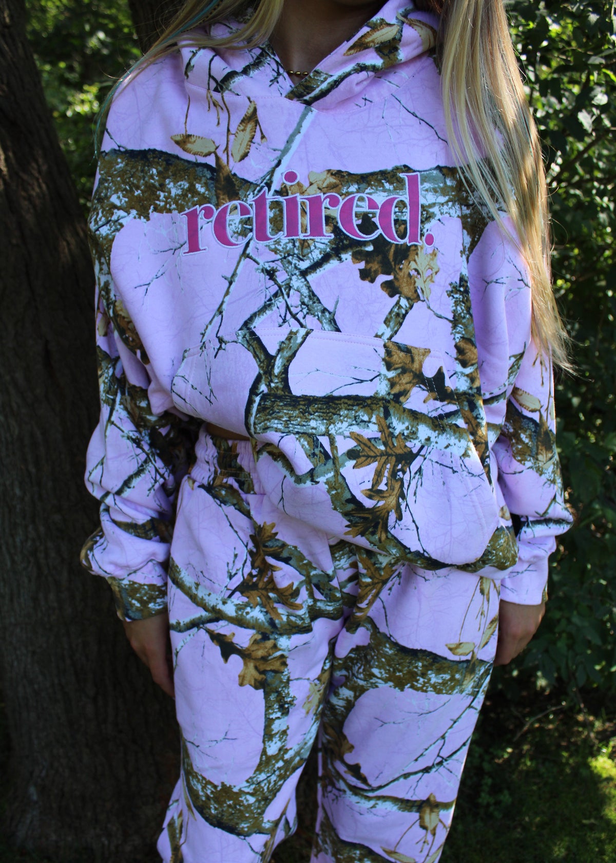 A person wearing a matching two-piece tracksuit with a pink and green woodland camouflage pattern, featuring the word 'retired.' printed in pink across the chest.