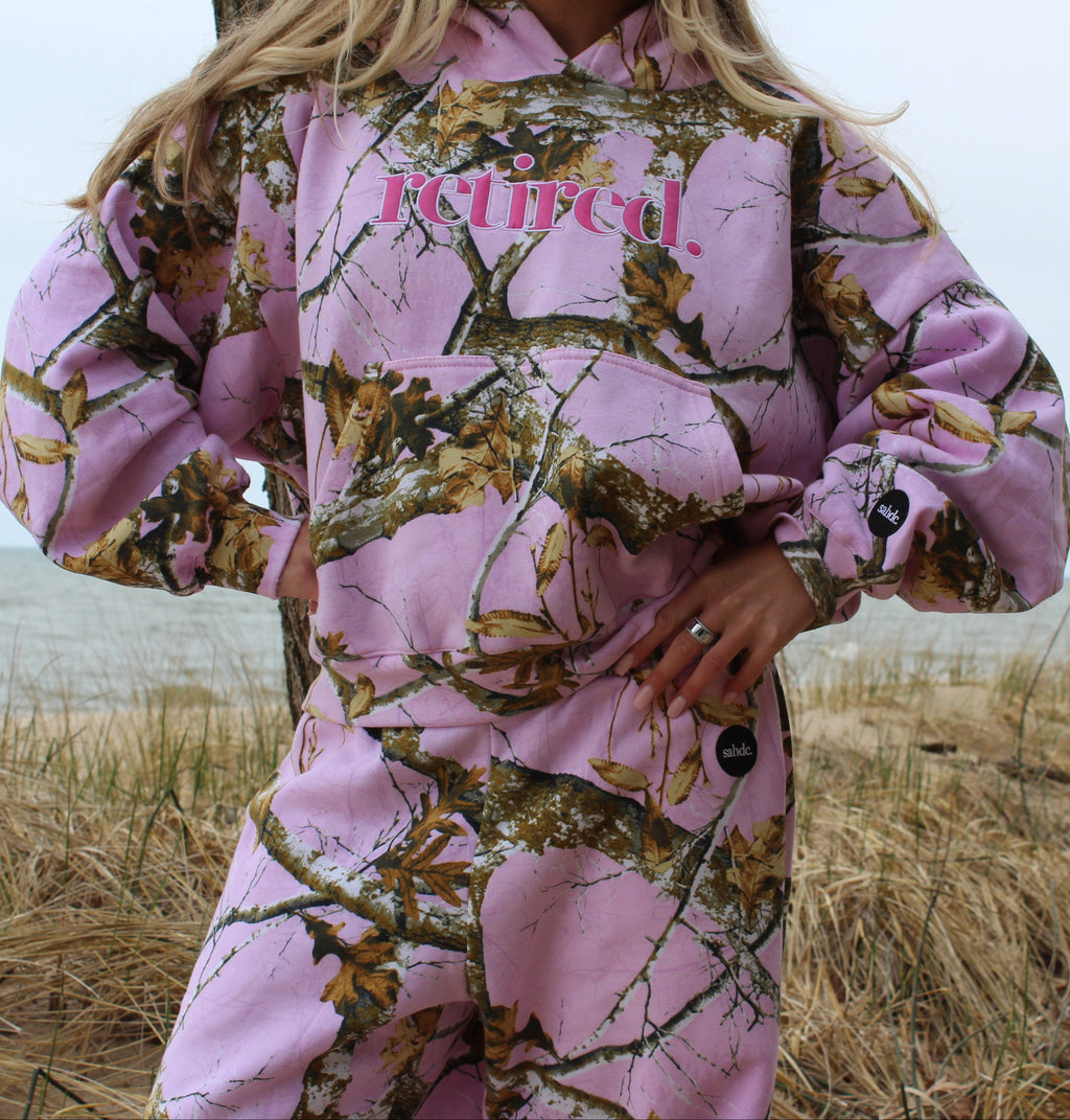 Person wearing a pink and camouflage hoodie with 'Retired' text,on the beach. 