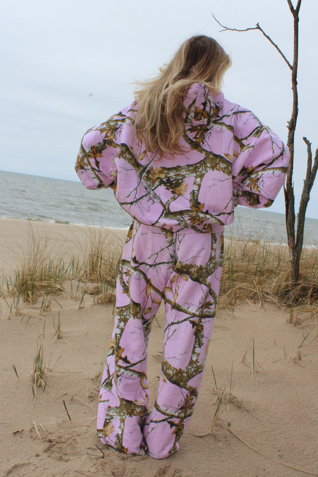 Person wearing a pink camouflage outfit standing on a sandy beach with a tree branch overhead.
