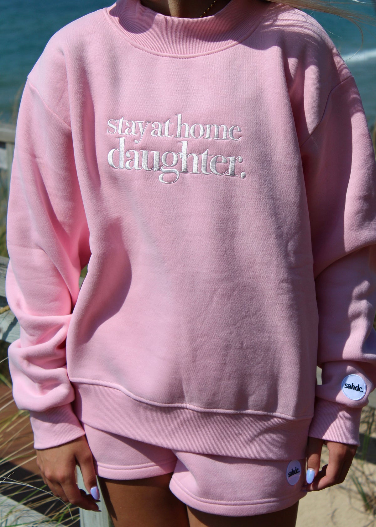 A person wearing a light pink crewneck sweatshirt with the phrase 'stay at home daughter.' in white script across the chest, paired with matching pink shorts, photographed outdoors near water and sand.