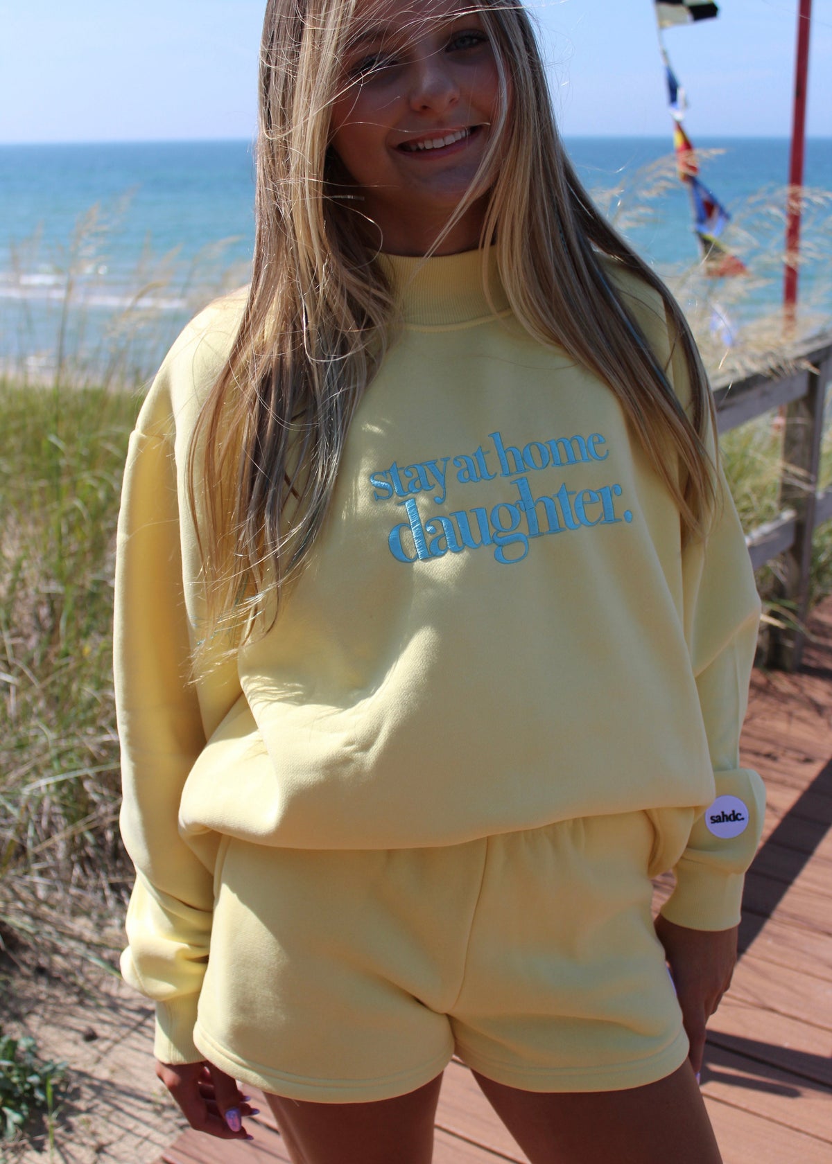 A woman wearing a pale yellow sweatshirt and matching shorts set, standing on a wooden boardwalk with the ocean and beach grass in the background. The sweatshirt features the text 'Stay at home daughter.