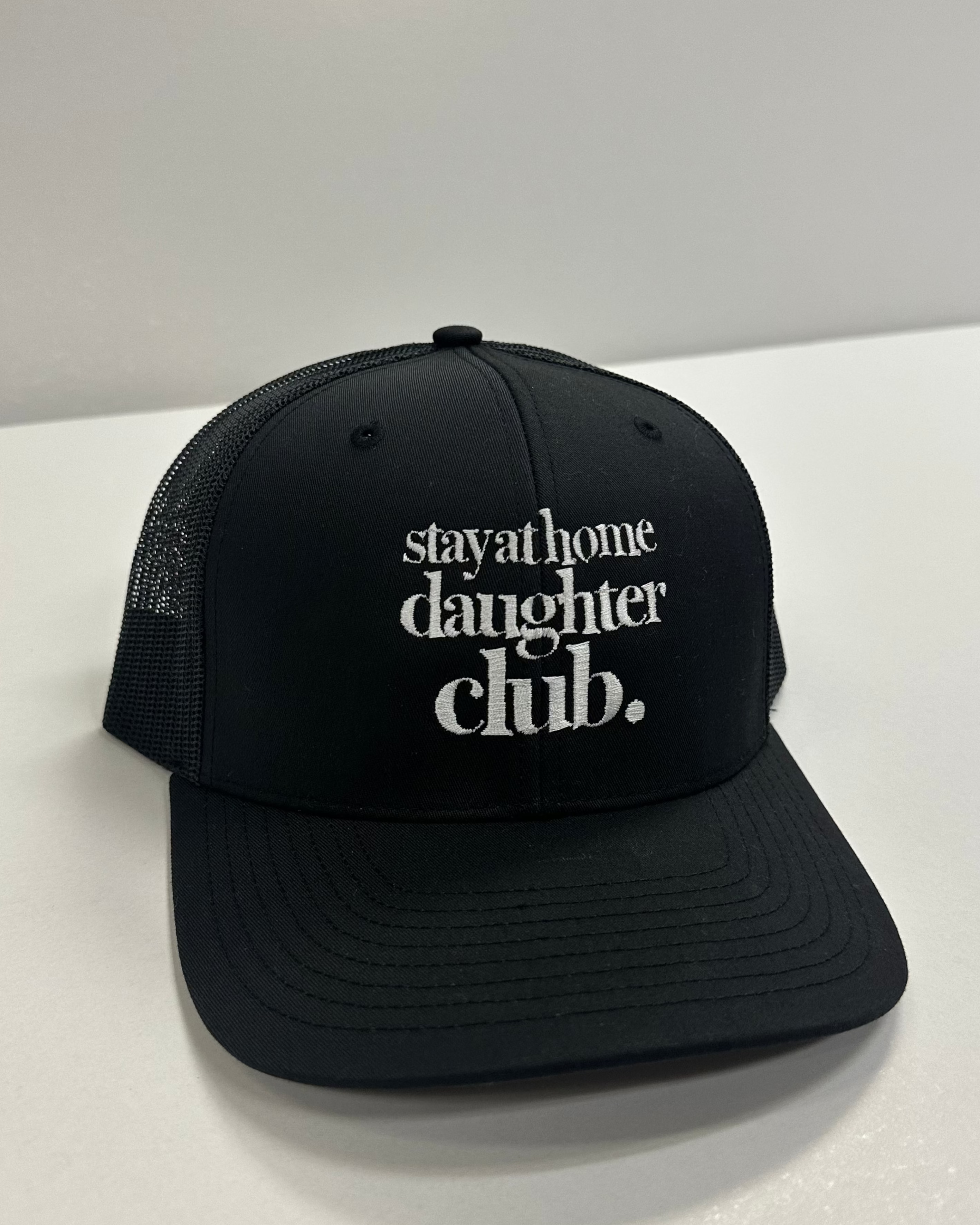 Black cap with 'stay at home daughter club' text on a white surface
