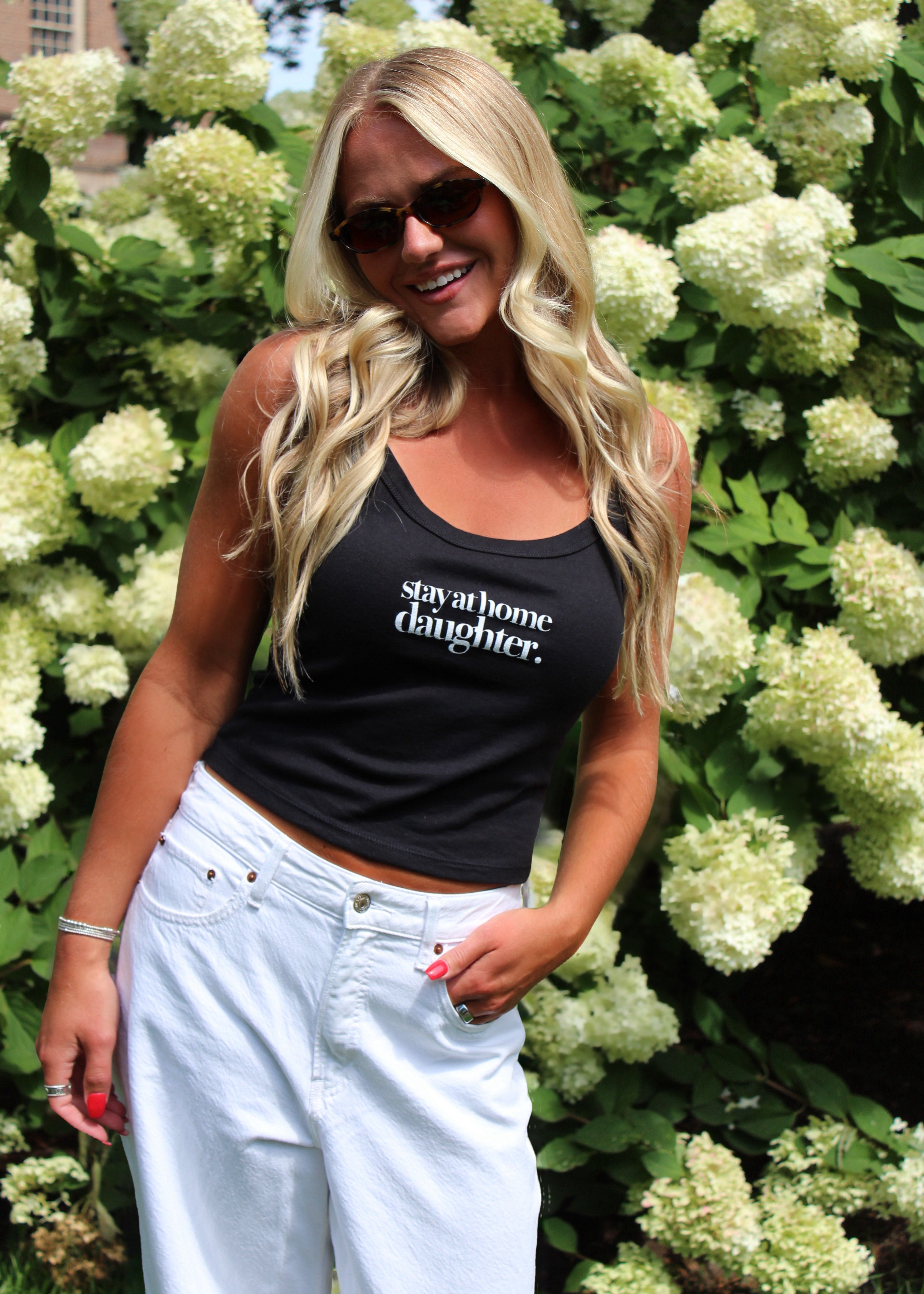 A blonde woman wearing a black tank top with the white text 'say it like a daughter.' and light-wash white jeans, standing outdoors in front of a dense background of white flowers and green foliage.