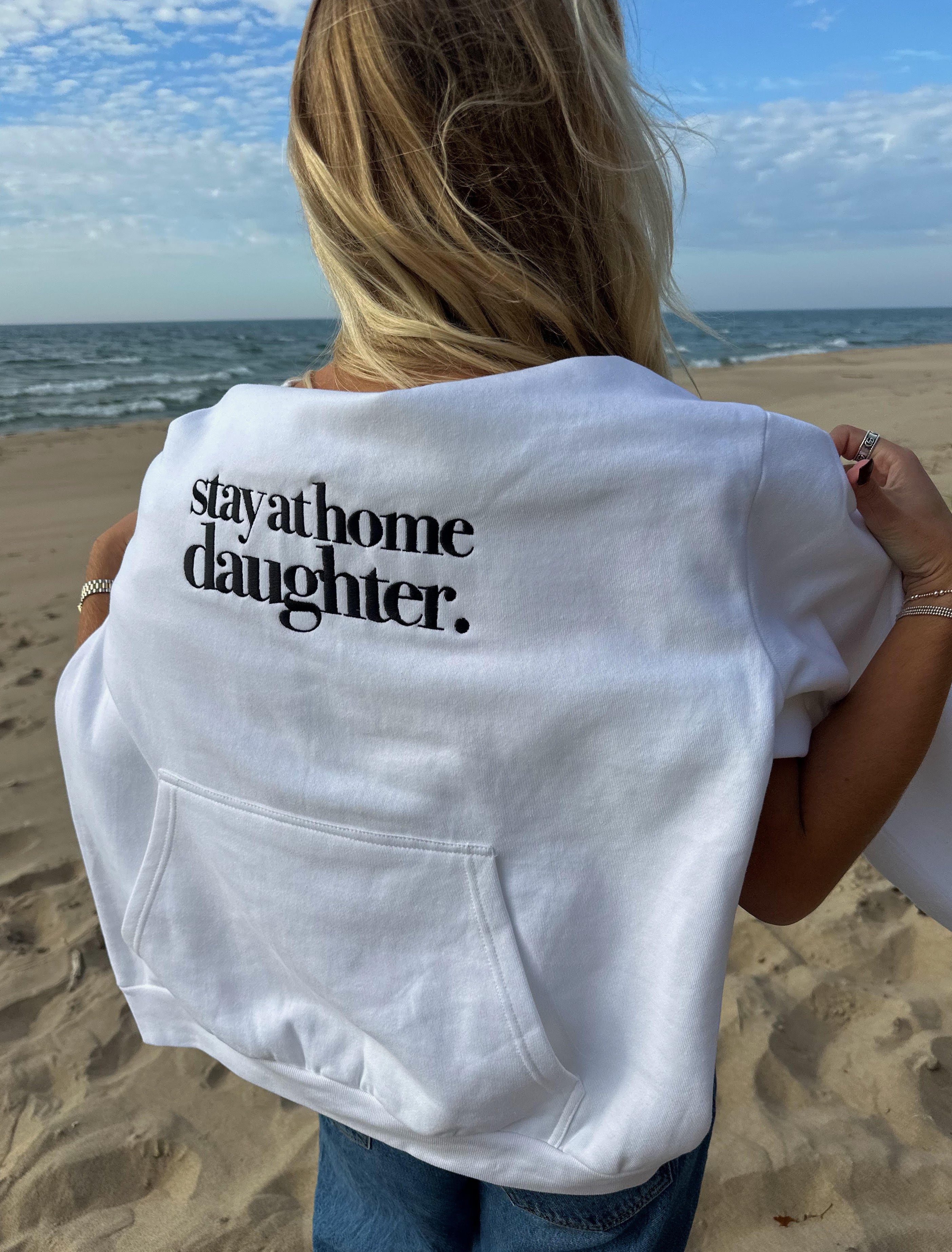 Person wearing a white sweatshirt with 'stay at home daughter' text on a beach