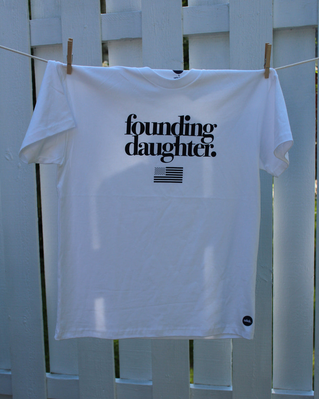 White t-shirt with 'founding daughter' text hanging on a clothesline against a white picket fence.