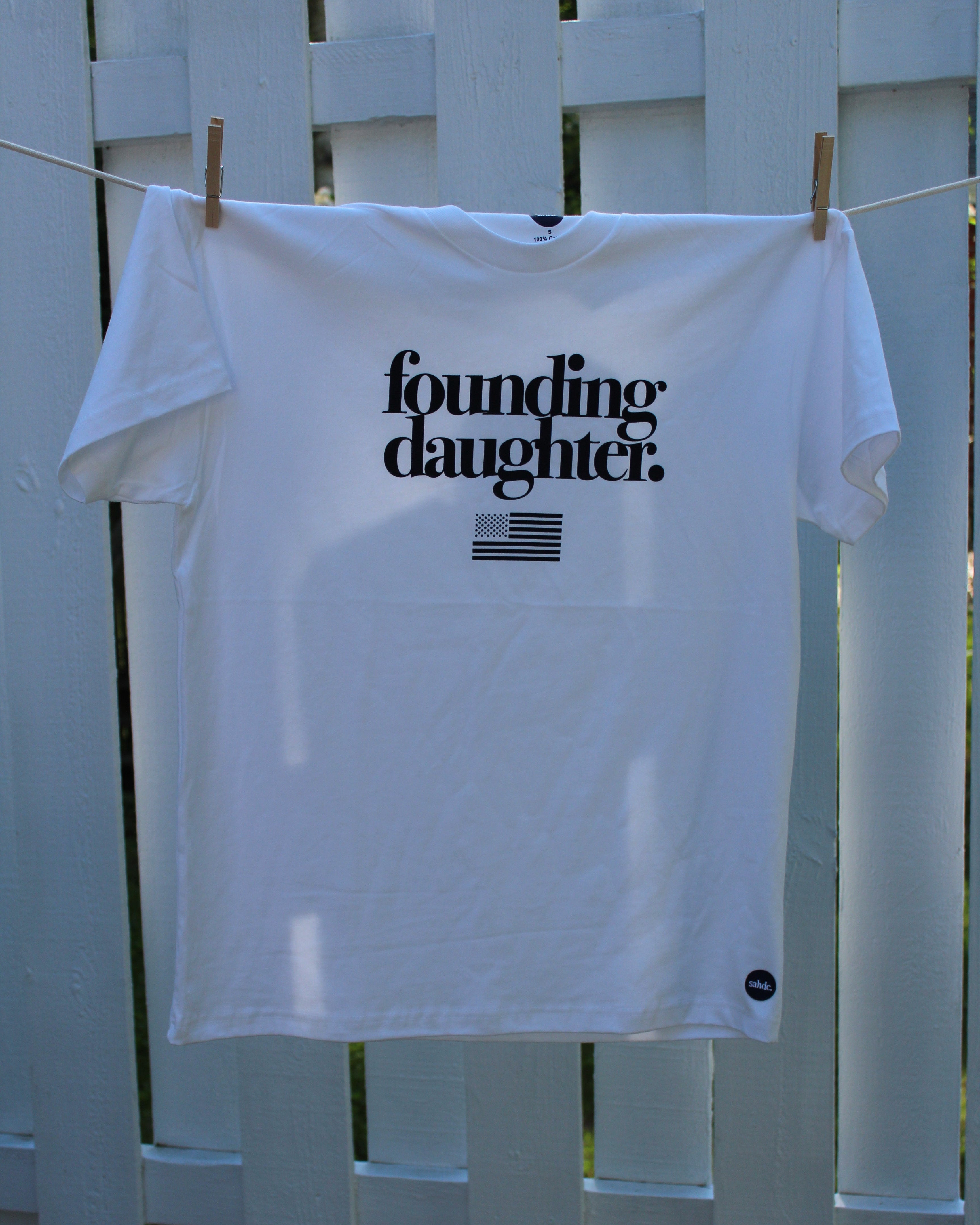 White t-shirt with 'founding daughter' text hanging on a clothesline against a white picket fence.