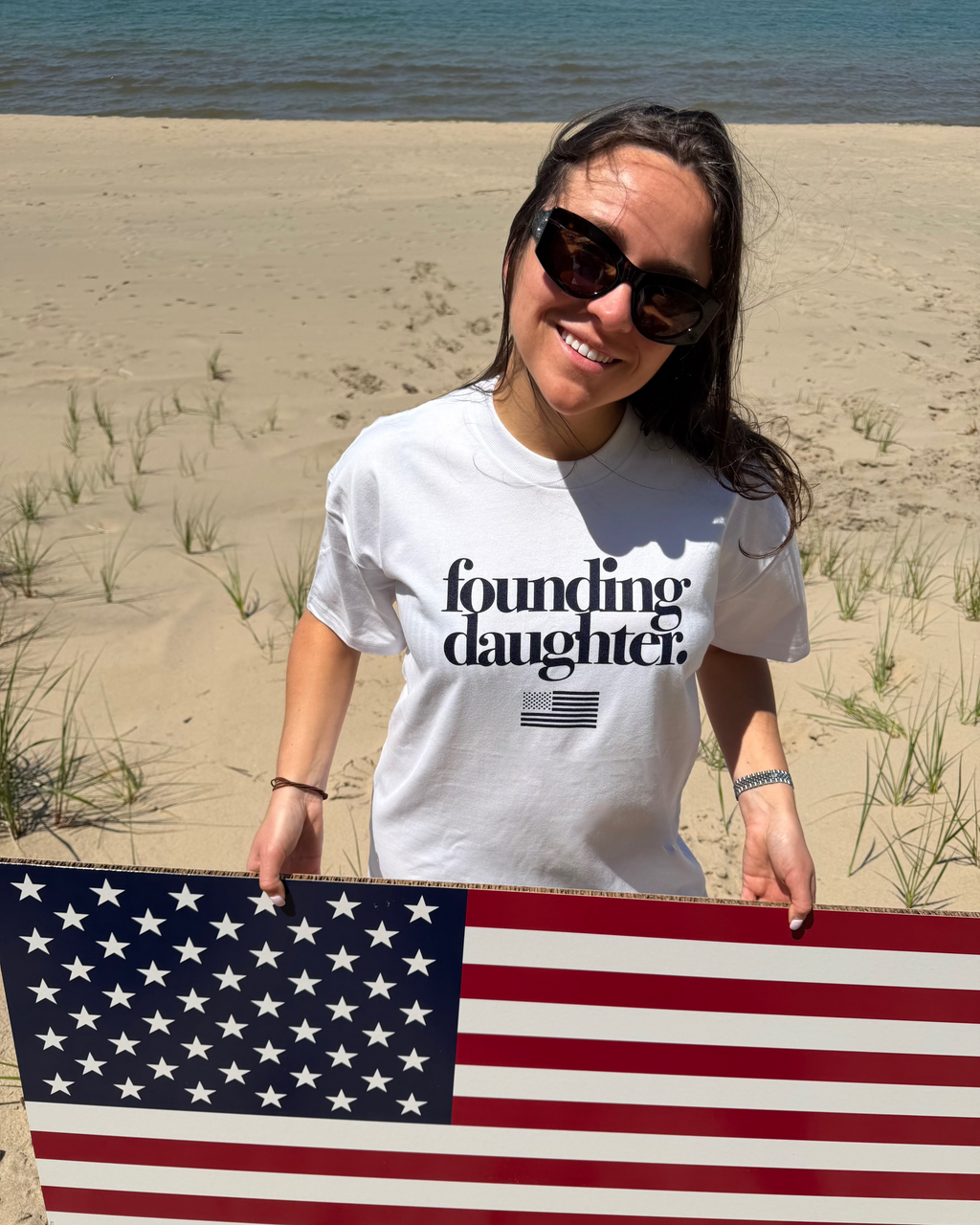 Woman holding an American flag on a beach wearing a 'foundling daughter' t-shirt.