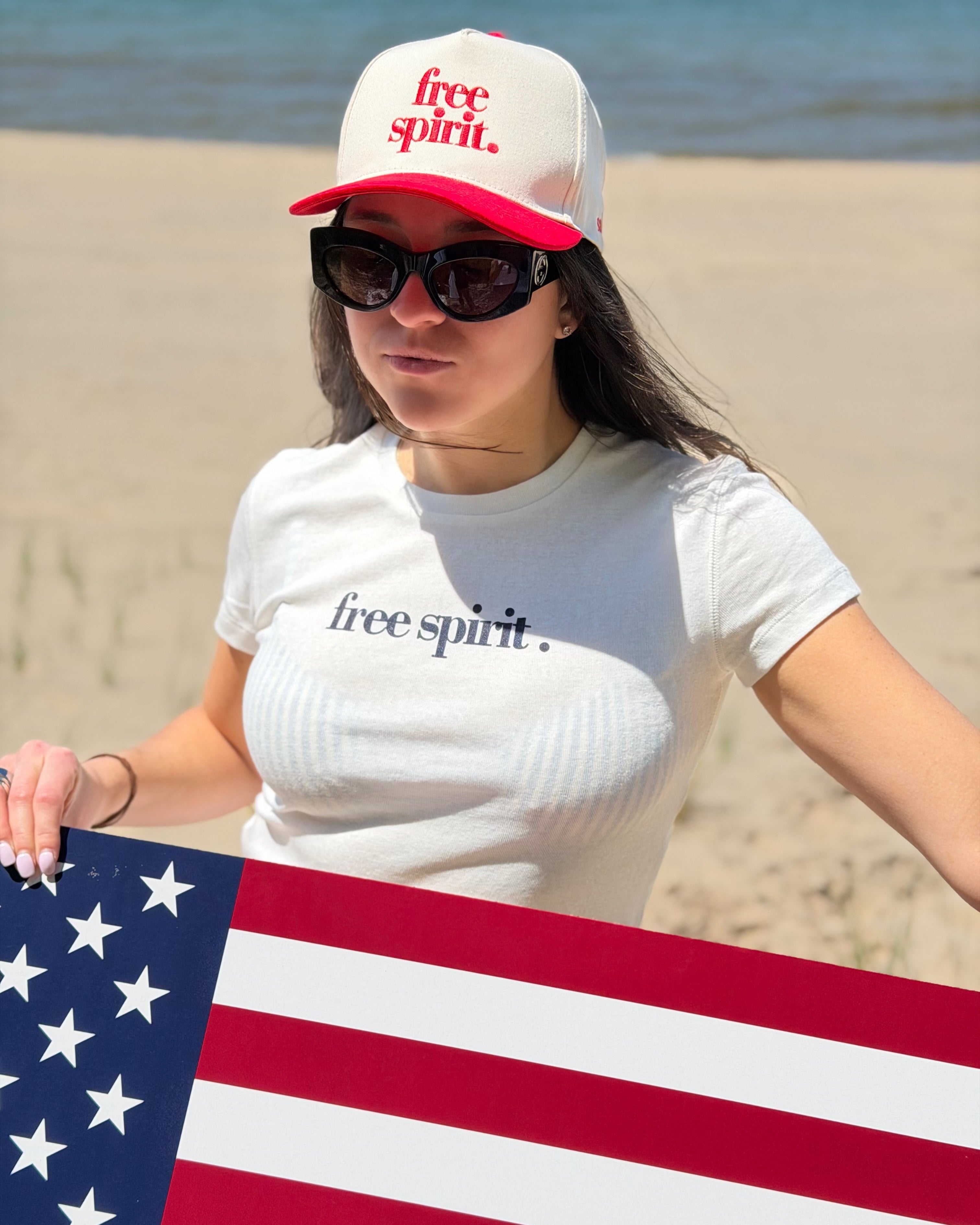 Woman wearing a 'free spirit' cap and t-shirt, holding an American flag on a beach.