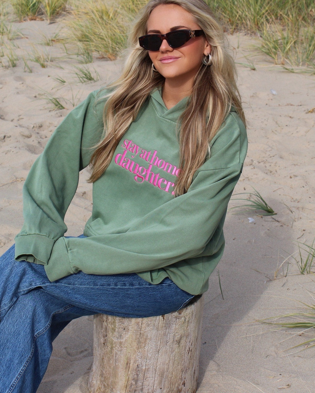 Woman sitting on a beach wearing a green sweatshirt with pink text and blue jeans.