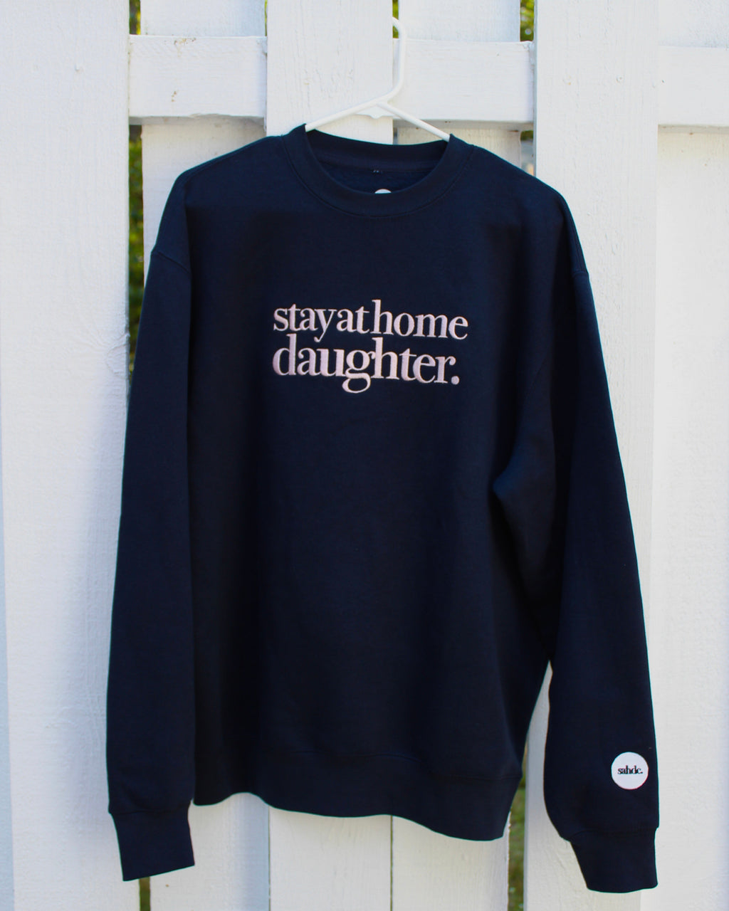 A navy blue crewneck sweatshirt hanging on a white hanger against a white wooden fence. The sweatshirt features the phrase 'stay at home daughter.