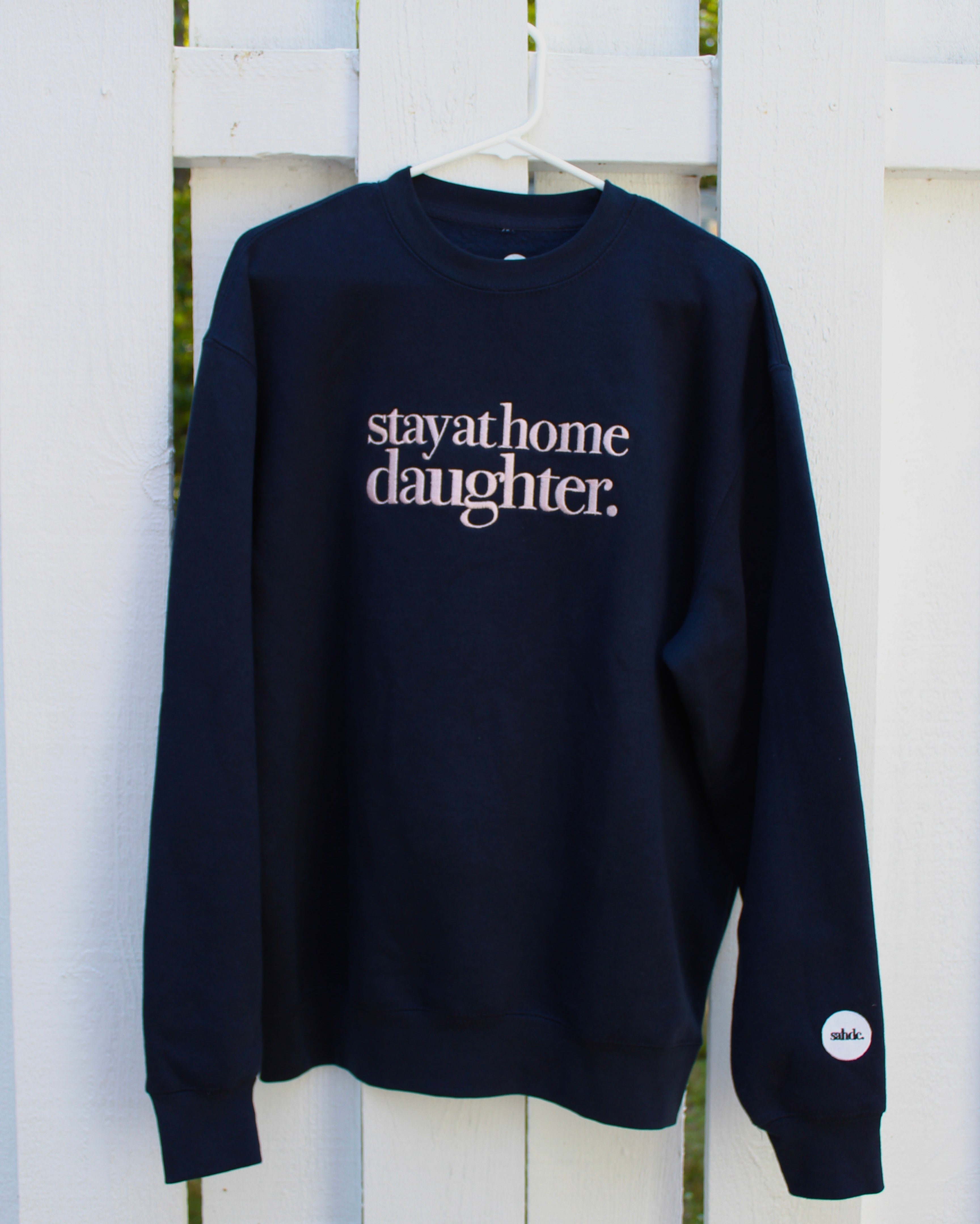 A navy blue crewneck sweatshirt hanging on a white hanger against a white wooden fence. The sweatshirt features the phrase 'stay at home daughter.