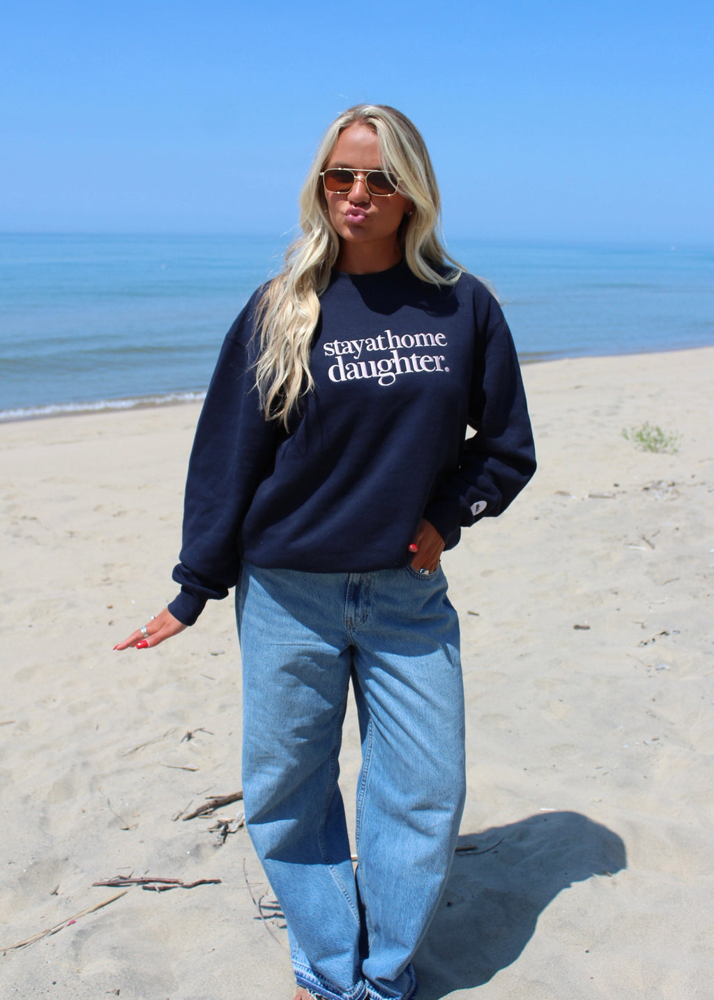 A woman stands on a sandy beach wearing a navy blue sweatshirt with the white text stay at home daughter and light blue jeans, with the ocean and blue sky in the background.