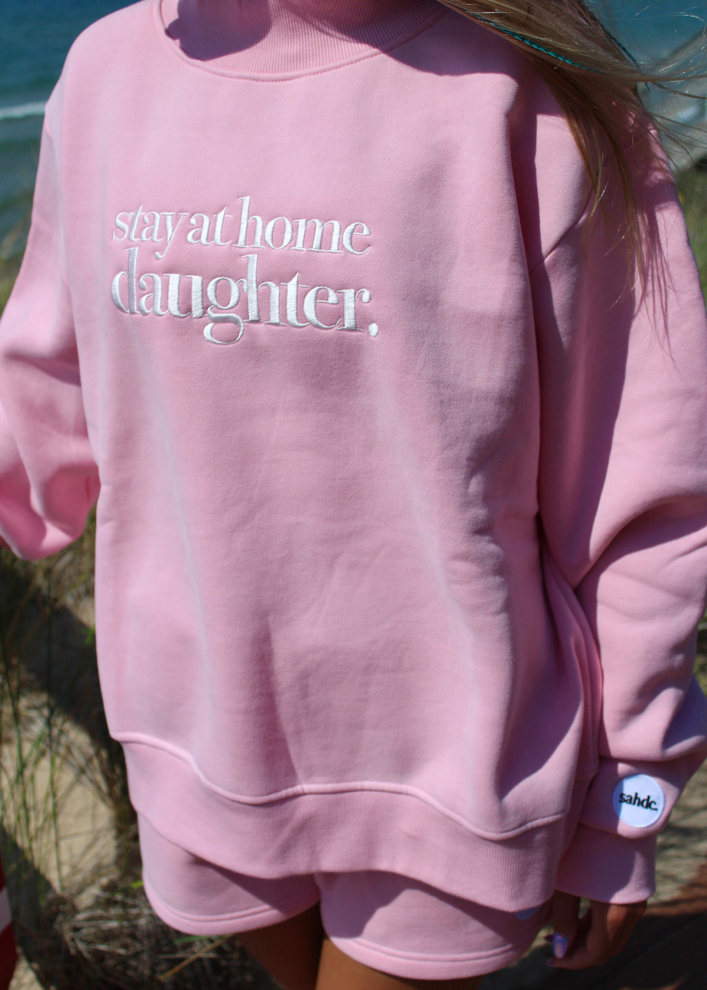 wearing a pink sweatshirt and matching shorts set outdoors near the beach. The sweatshirt has the words 'stay at home daughter,' printed in white across the chest