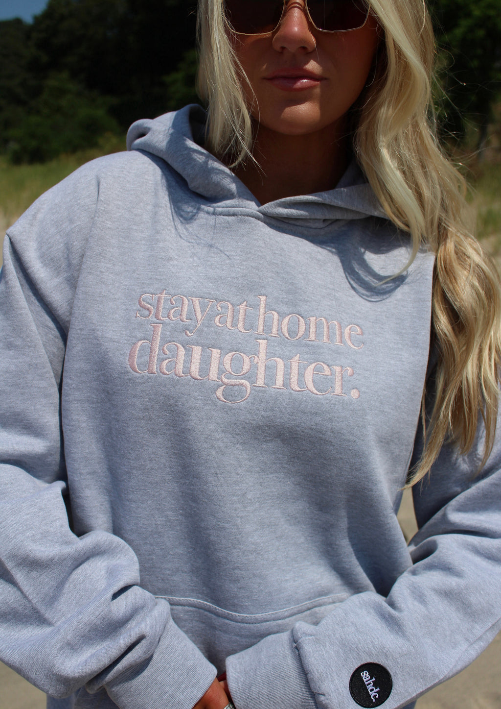A woman wearing a light heather gray hoodie with the words 'stay at home daughter.' printed in a dusty pink script across the chest.