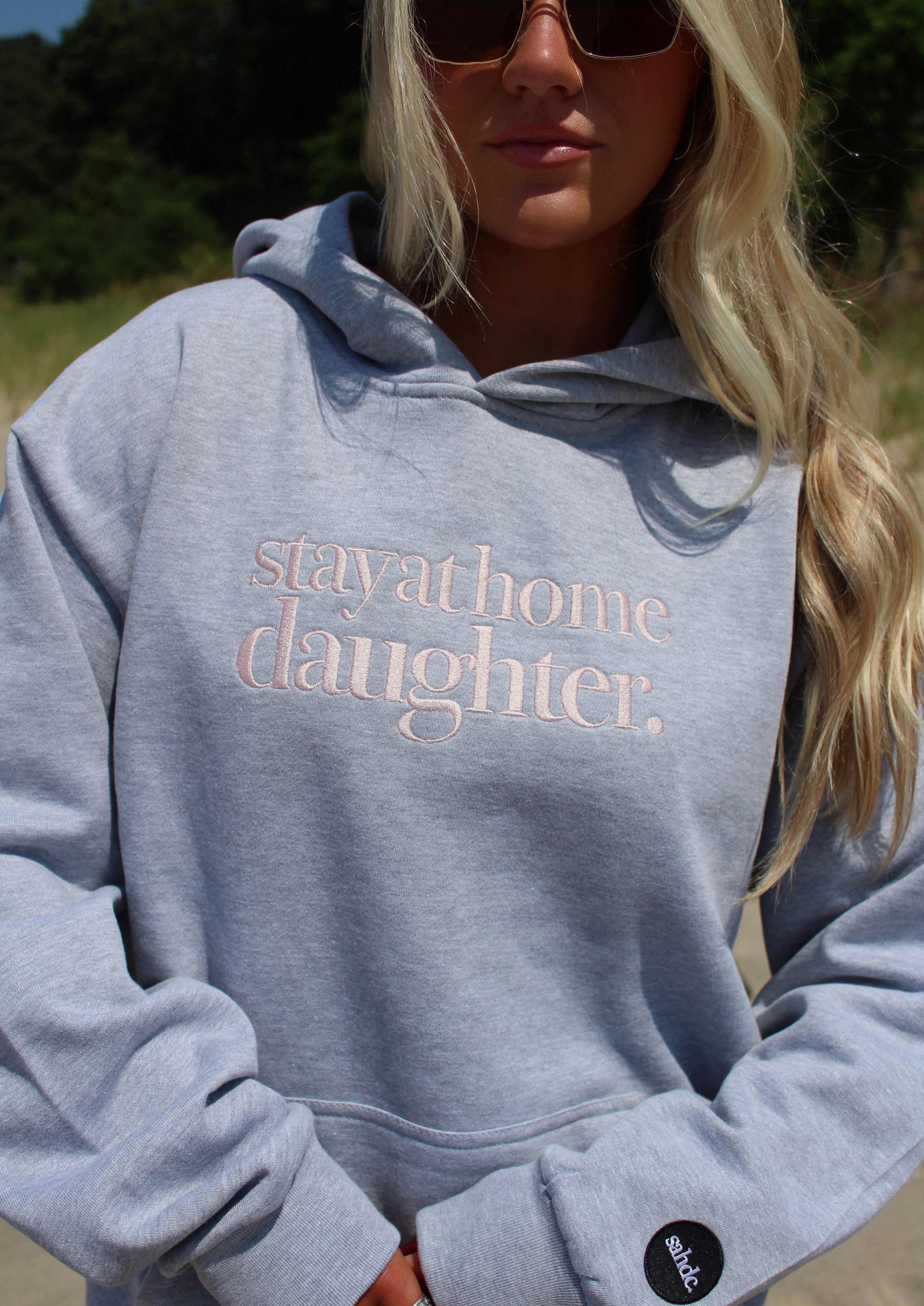 A woman wearing a light heather gray hoodie with the words 'stay at home daughter.' printed in a dusty pink script across the chest.