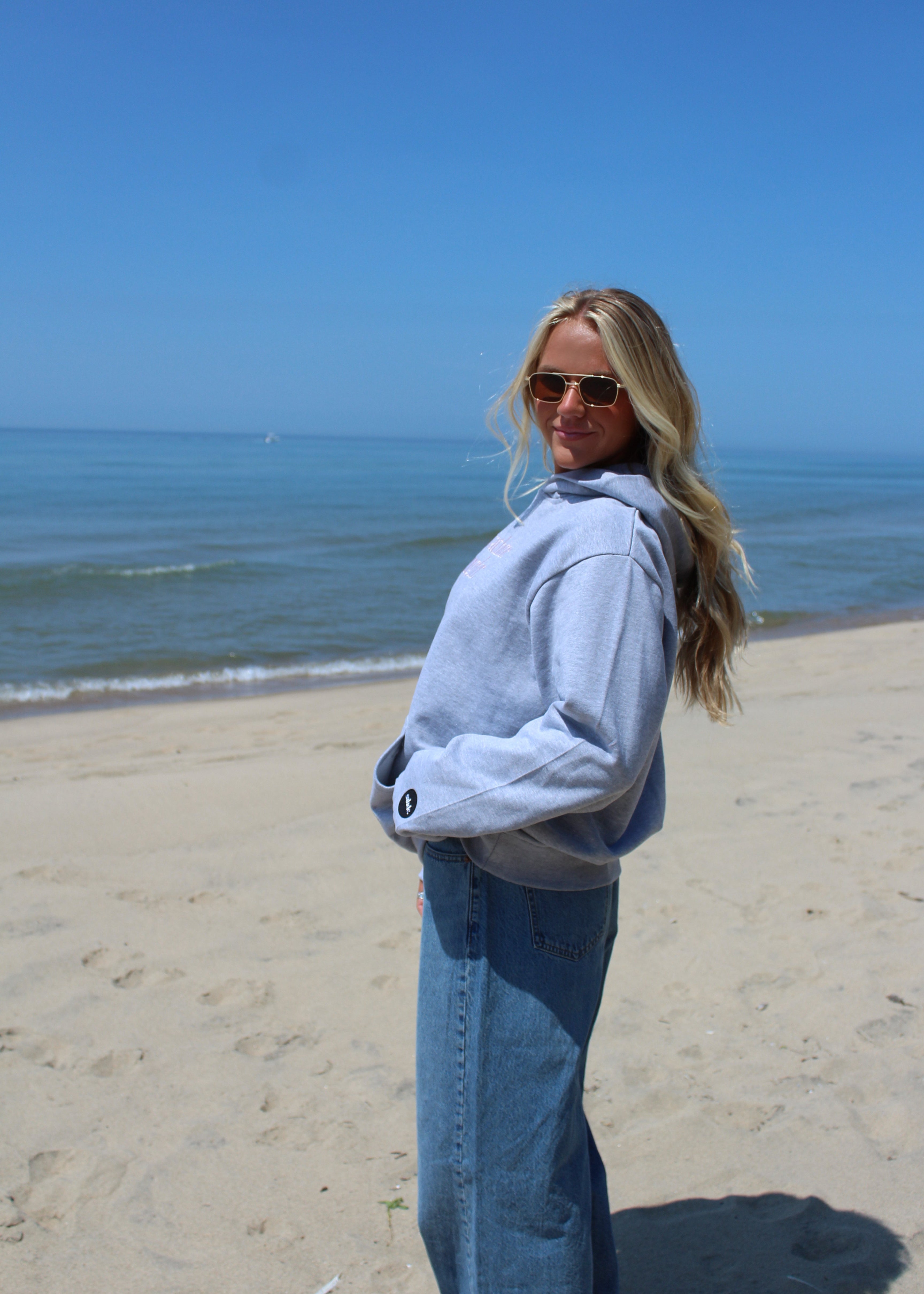 A woman with blonde hair wearing a light gray oversized hoodie and blue jeans stands on a sandy beach with the ocean and blue sky in the background.