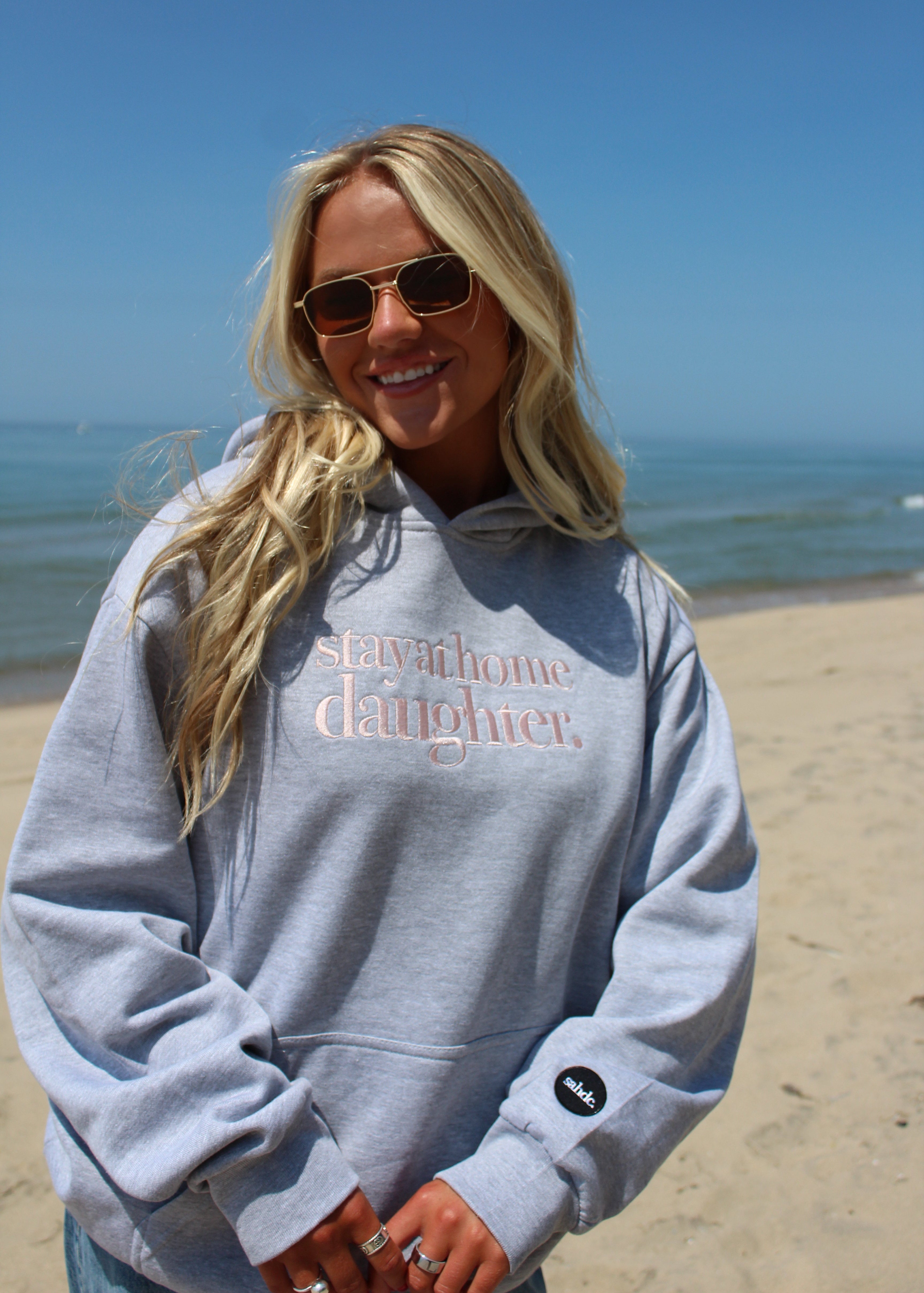 A woman wearing a light gray hoodie with stay at home daughter printed in pink on the chest, standing on a sandy beach with the ocean and blue sky in the background.