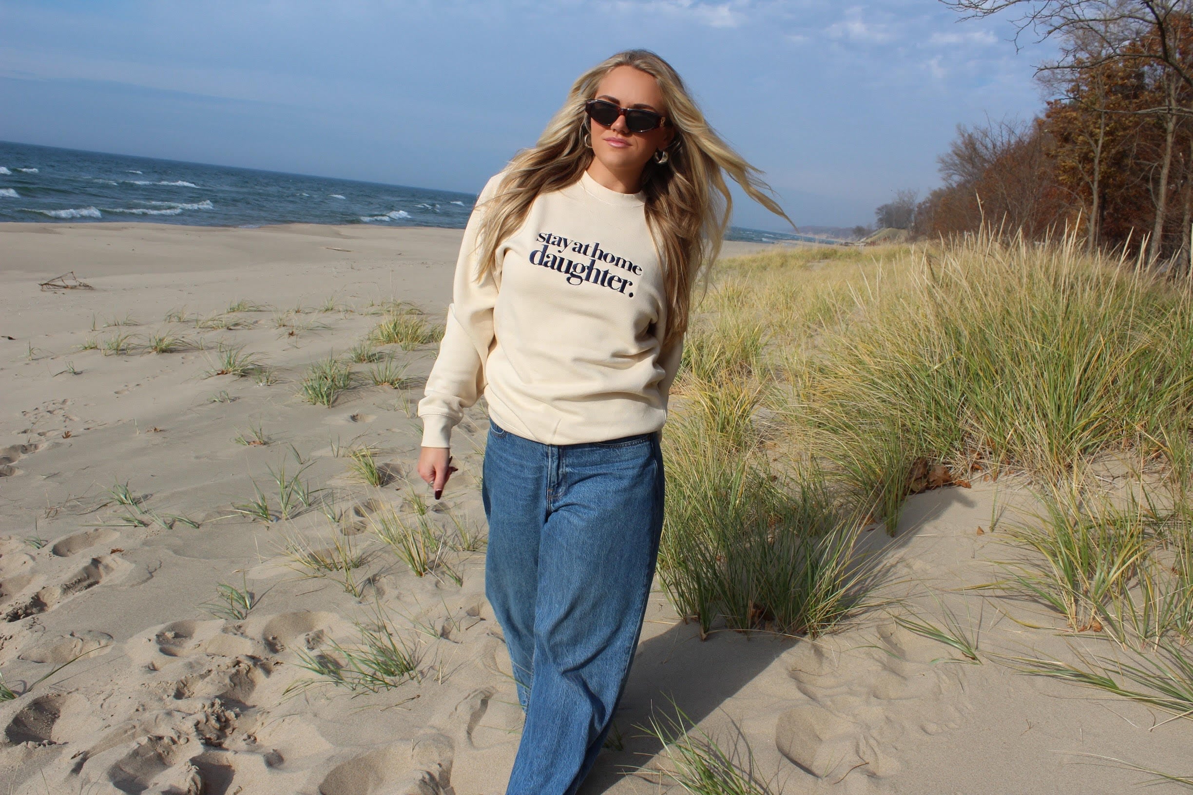 Woman walking on a sandy beach with grasses, wearing a beige sweatshirt and sunglasses.