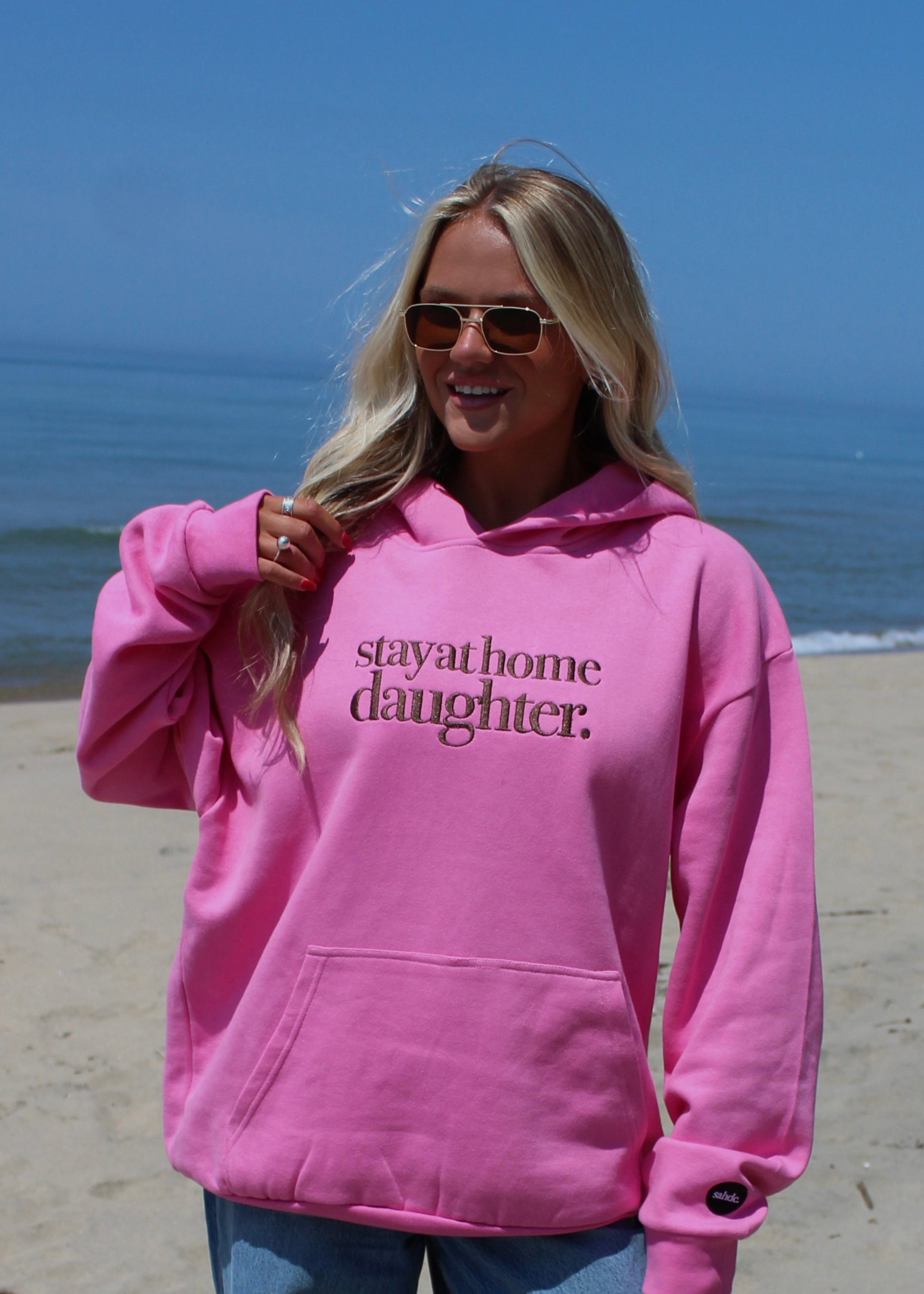 A woman wearing a bright pink hoodie with the text 'stay at home daughter.' printed in dark brown across the chest, standing on a sandy beach with the ocean and blue sky in the background.